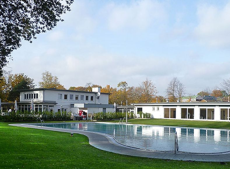 Fotografie: Außenaufnahme; Blick über gepflegte Rasenfläche auf flachen weißen Bau mit großen Fensterflächen; davor ein natürlich geformter Pool; umliegende Bäume