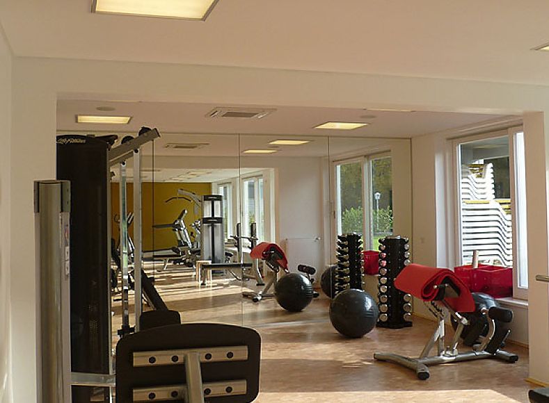 Fotografie: Innenaufnahme; moderner Fitnessraum mit Kraftgeräten, Hanteln und Spiegelwand; große Fenster sorgen für helle, offene Atmosphäre; heller Fußboden