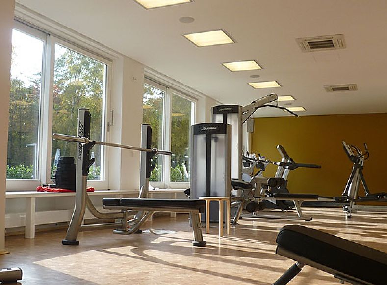 Fotografie: Trainingsraum mit verschiedenen Fitnessgeräten; große Fensterfronten und Deckenspots sorgen für helle Raumatmosphäre; freie Flächen zwischen Geräten betonen Offenheit